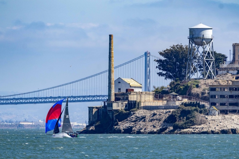 Sailboat with a red and blue spinnaker sailing by Alcatraz with the Bay Bridge behind